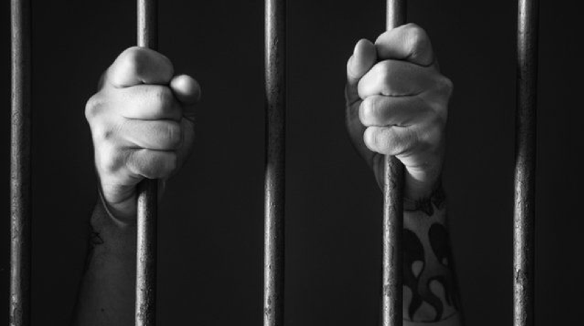 black and white photo of a person's hands and forearms grabbing prison bars