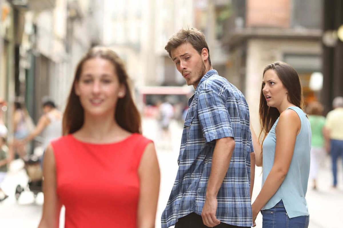 Color photo of the meme depicting a man holding hands with a woman, but the man is looking behind him lustfully at another woman walking the other way while the woman he is holding hands with gives him a look of disgust